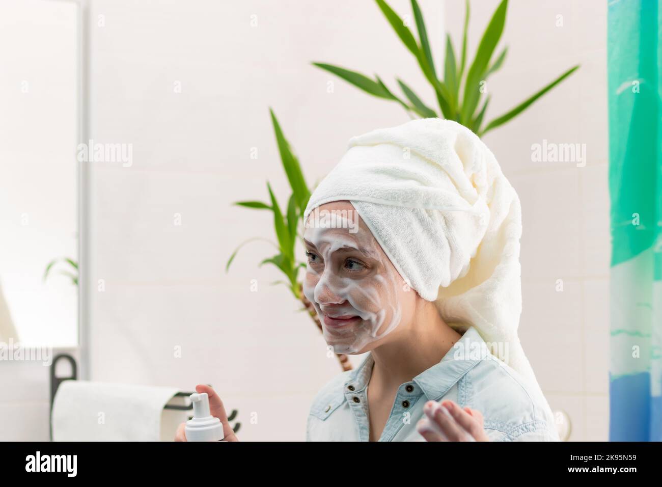 Una giovane bella donna con un asciugamano bianco sulla testa in bagno applicato trucco Rimuovi alla sua faccia contro lo sfondo di una pianta verde. SEL Foto Stock