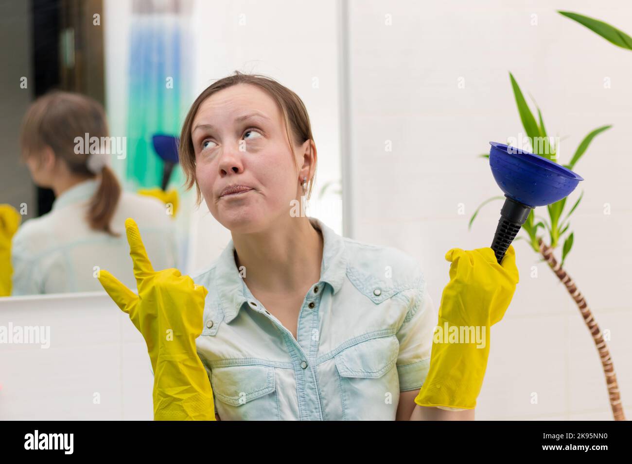 Una giovane donna pulitore in giallo guanti di gomma sanitaria in un bagno luminoso con uno stantuffo per eliminare il blocco nelle sue mani. Messa a fuoco selettiva. PORTRA Foto Stock