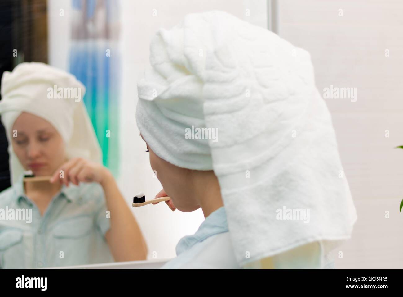 Una donna giovane e graziosa con un asciugamano sulla testa in un bagno luminoso con uno spazzolino in mano le spazzolerà i denti. Riflessione nello specchio. Selectiv Foto Stock