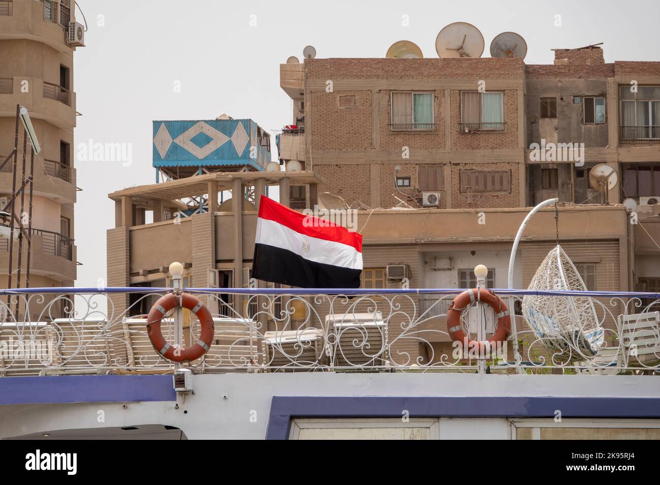 La bandiera egiziana che vola dal ponte superiore di una nave da crociera ormeggiata sul Nilo con edifici di appartamenti sullo sfondo Foto Stock