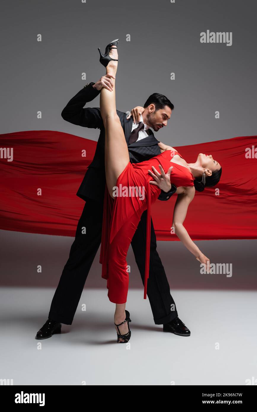 Vista laterale di un'elegante ballerina che tocca la gamba del partner mentre si esegue il tango su sfondo grigio con tessuto rosso Foto Stock