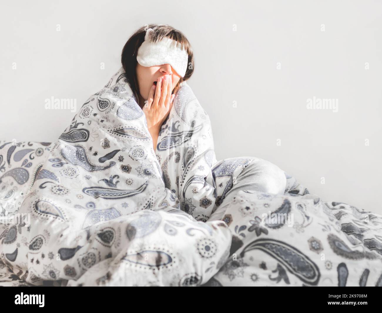 Donna in pajama grigia e maschera del sonno sul viso. Si è appena svegliata e si siede a letto. Mattina presto in casa accogliente. Foto Stock