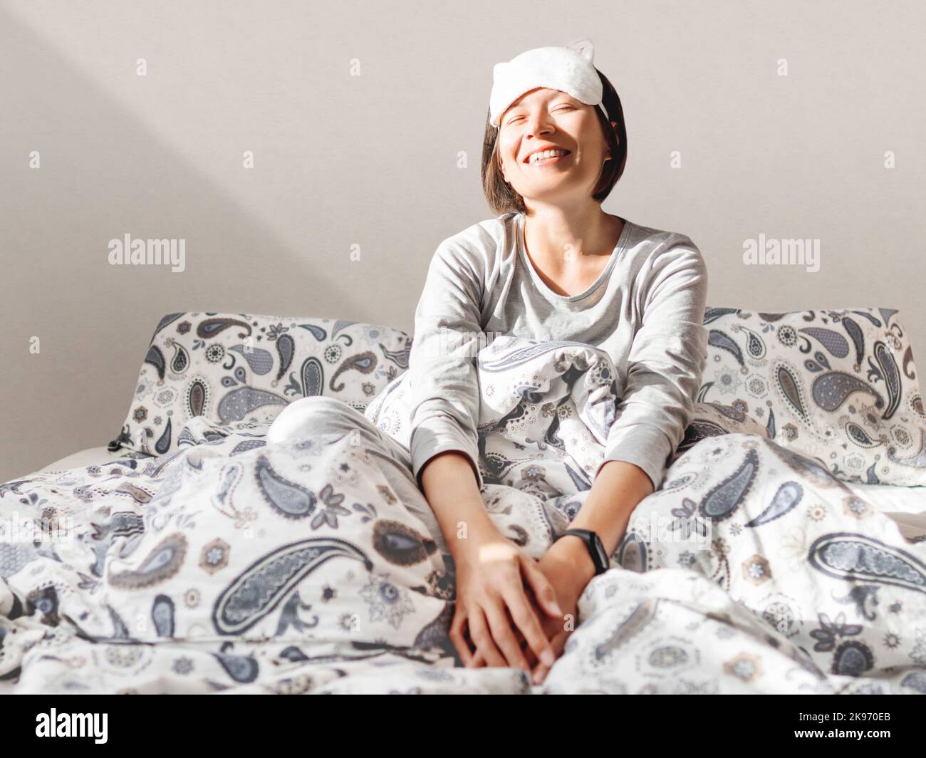 Donna sorridente in pajama grigio e maschera del sonno. Si è appena svegliata e si siede a letto. Mattina presto in casa accogliente. Foto Stock