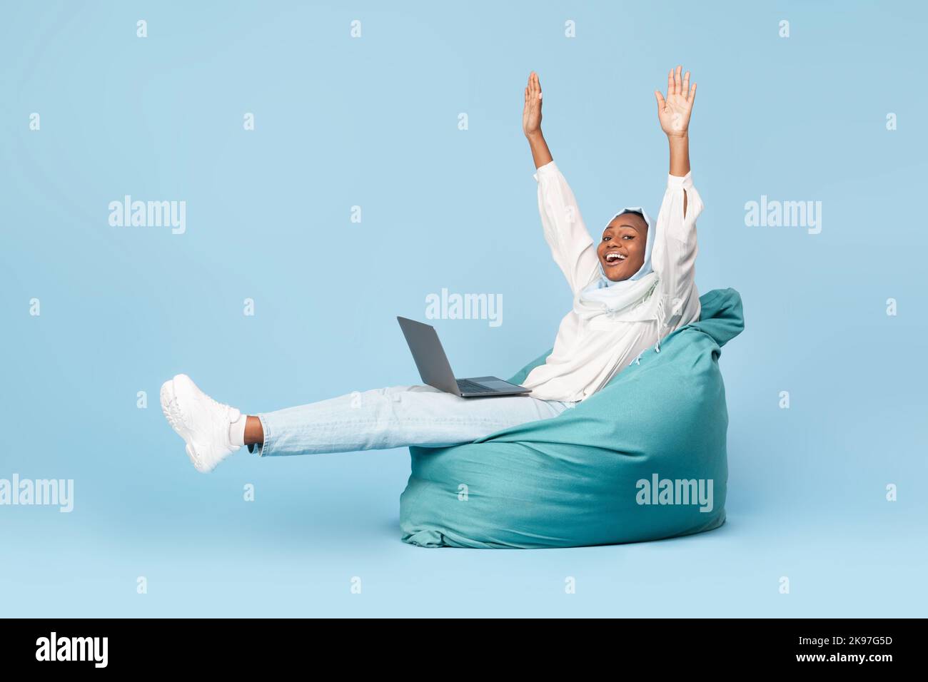 Donna musulmana afro-americana overjoyed seduta in sedia di beanbag con il laptop e alzando le mani in su, ha ottenuto le notizie grandi Foto Stock