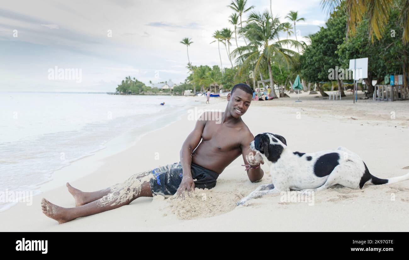 Buon turista afro-americano maschio in pantaloncini seduti sulla sabbia e accarezzare cane obbediente mentre si passa il fine settimana estivo giorno in spiaggia Foto Stock