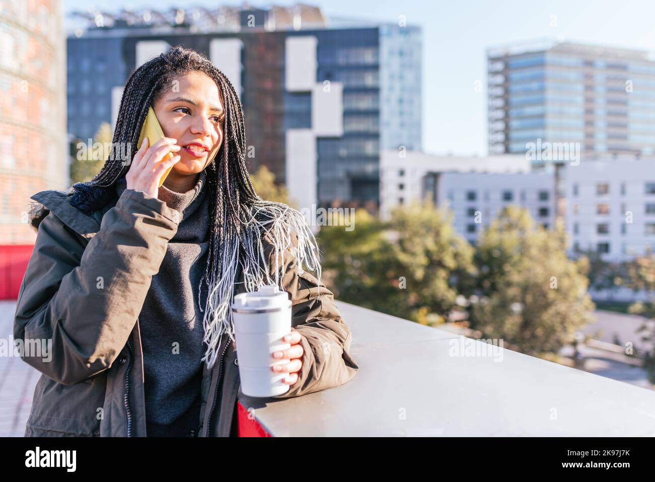 Contenuto femmina ispanica con Dreadlocks che hanno una telefonata mentre in piedi vicino al confine con la coppa termica in città con edifici residenziali Foto Stock