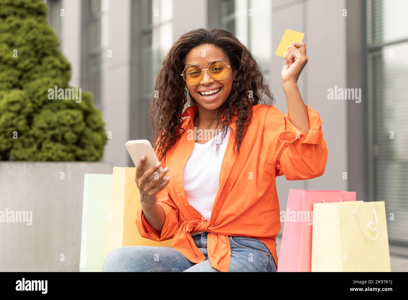 Ridendo millenario afroamericana donna in occhiali con molti sacchetti mostrare carta di credito e digitando sullo smartphone Foto Stock