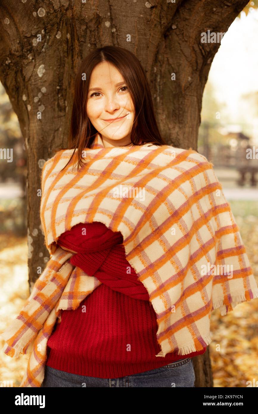 Ritratto femminile di felice bella donna in confortevole maglione rosso lavorato a maglia coperto con scacchi caldo coperta, albero tronco sfondo. Giovane donna entrare Foto Stock