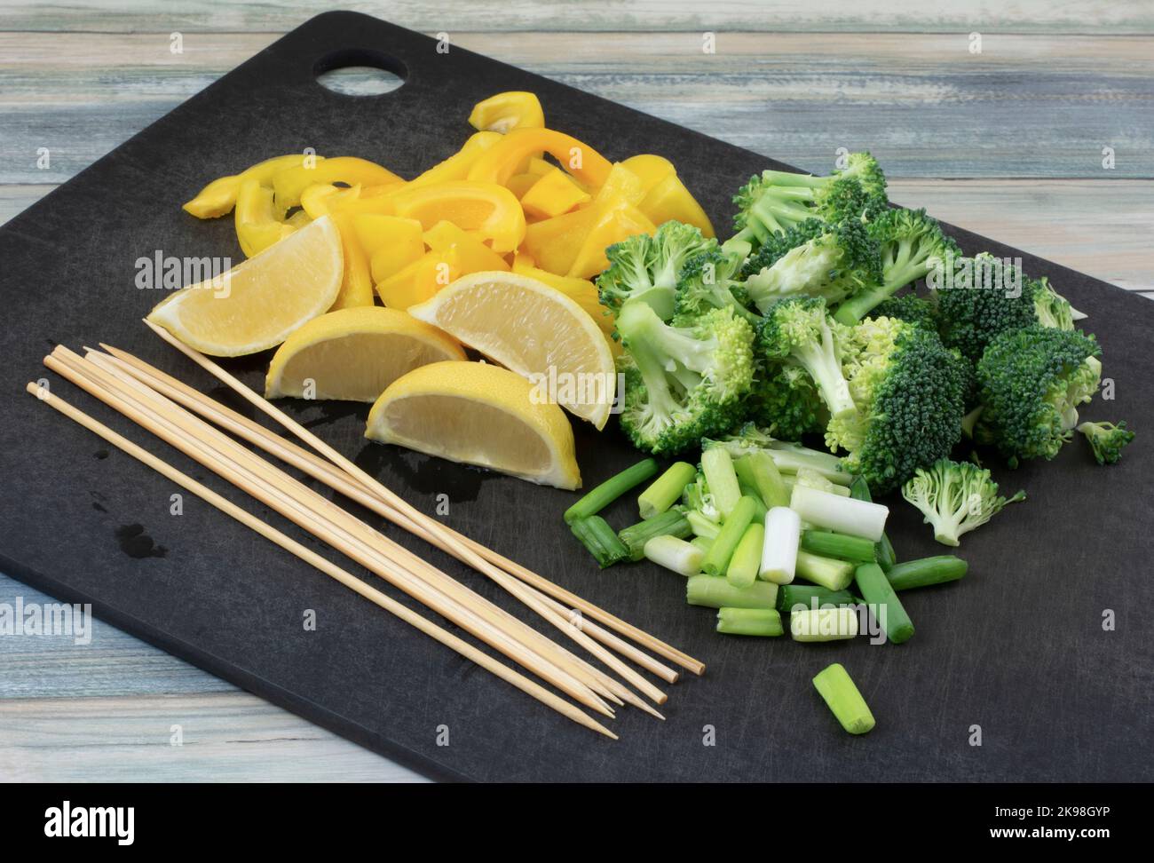 Broccoli tritati e cipolle verdi tagliate a fette, limone e peperone giallo con spiedini sul tagliere nero Foto Stock