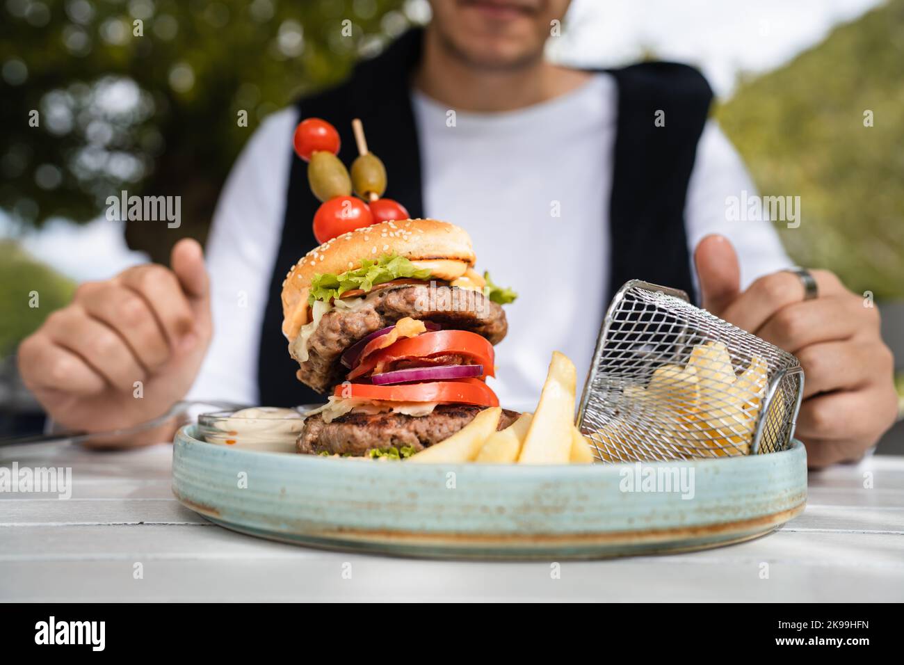 primo piano su un hamburger in un piatto e le mani di sconosciuto uomo caucasico prepararsi a mangiare sul tavolo nella giornata di sole all'aperto al ristorante Foto Stock