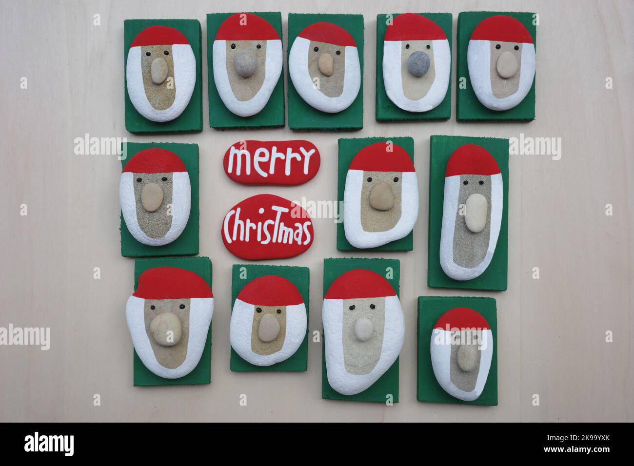 Buon messaggio natalizio composto da teste di Babbo Natale fatte a mano con pietre colorate dipinte a mano e ciottoli su un asse di legno Foto Stock