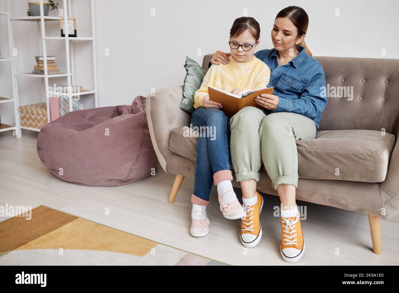 Ritratto completo di ragazza adolescente con sindrome di Down lettura libro con la madre amorevole a casa Foto Stock