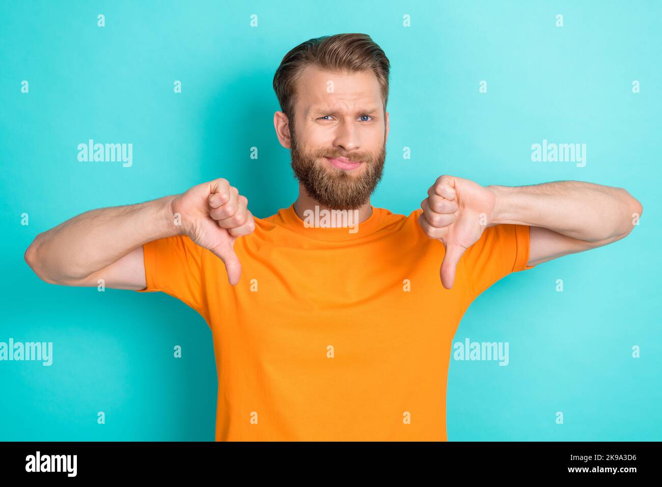 Ritratto di triste ragazzo infelice con barba bionda indossare t-shirt arancione mostrando pollici giù antipatico isolato su sfondo colore teal Foto Stock