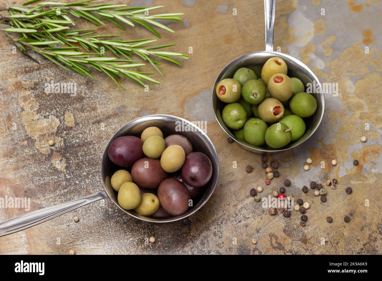 Olive di diverse varietà in una ciotola di metallo. Rosmarino e allspice. Giacitura piatta. Fondo in metallo arrugginito. Foto Stock