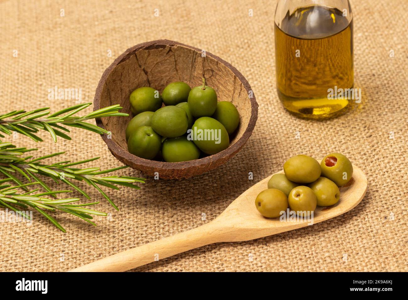 Olive verdi in cucchiaio di legno e in guscio di cocco. Olio d'oliva in bottiglia e un rametto di rosmarino. Vista dall'alto. Sfondo burlap Foto Stock