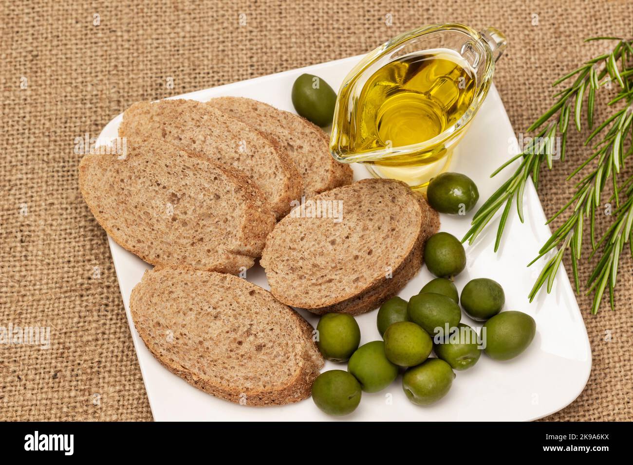 Olive verdi, fette di pane e olio in ciotola di vetro in piatto bianco. Rametto di rosmarino. Giacitura piatta. Sfondo burlap Foto Stock