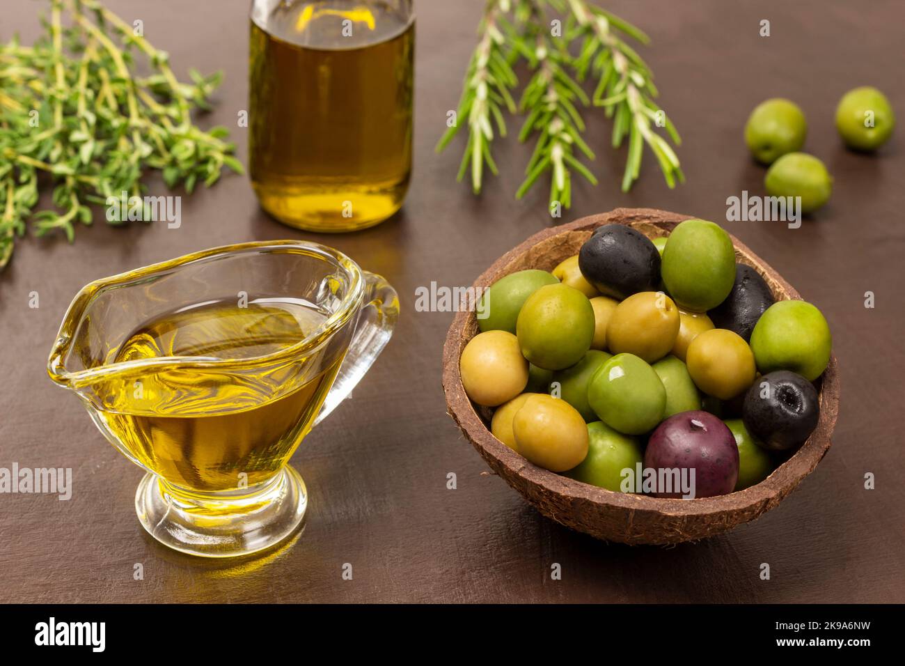 Olive di varie varietà in conchiglia di cocco. Olio d'oliva in ciotola e bottiglia. Ciuffi di timo e rosmarino. Vista dall'alto. Sfondo marrone. Foto Stock