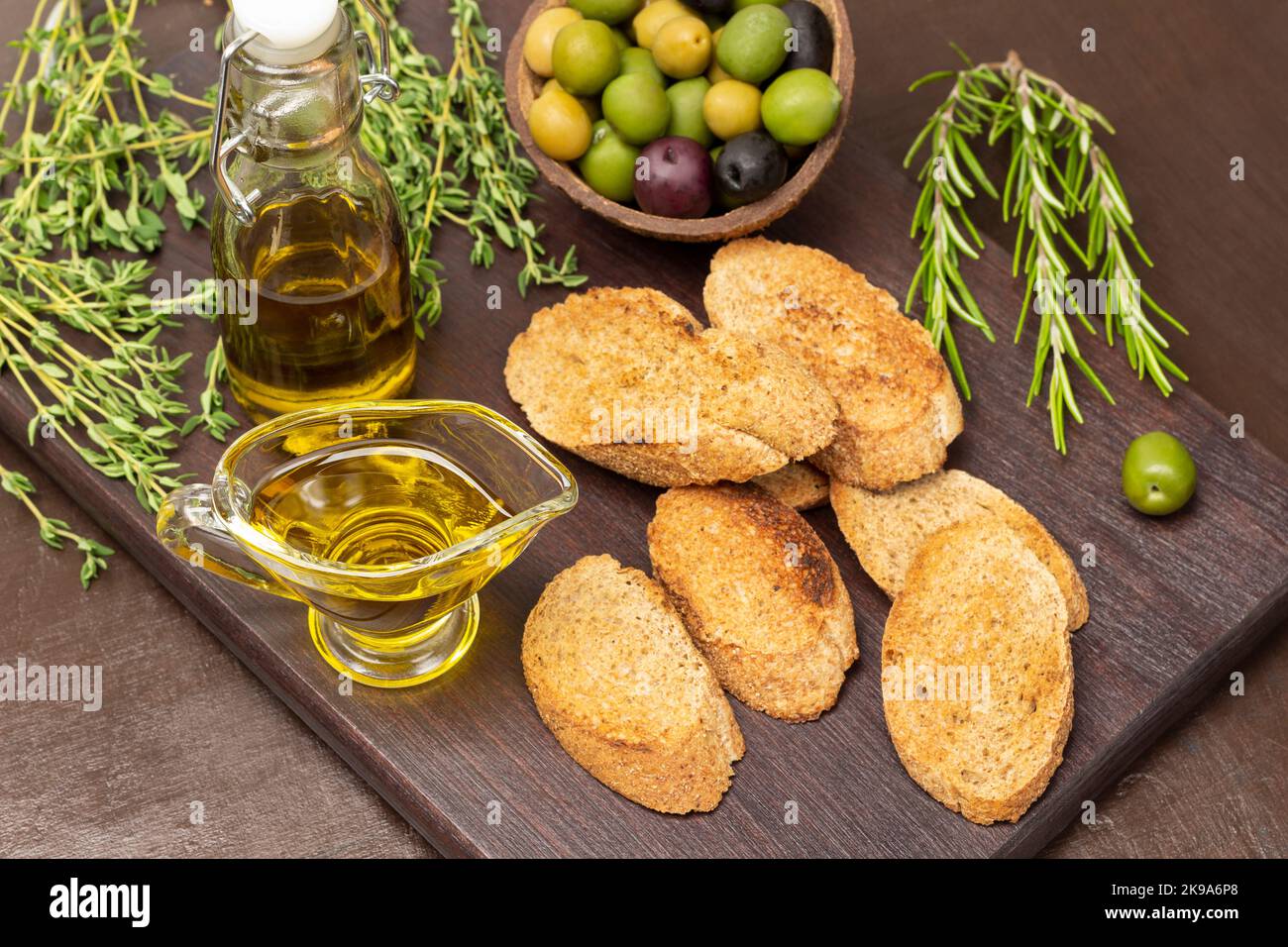 Fette di pane tostate, olive di varie varietà in conchiglia di cocco, rametti di timo e rosmarino, olio d'oliva in ciotola e bottiglia sul tagliere. Da a. Foto Stock