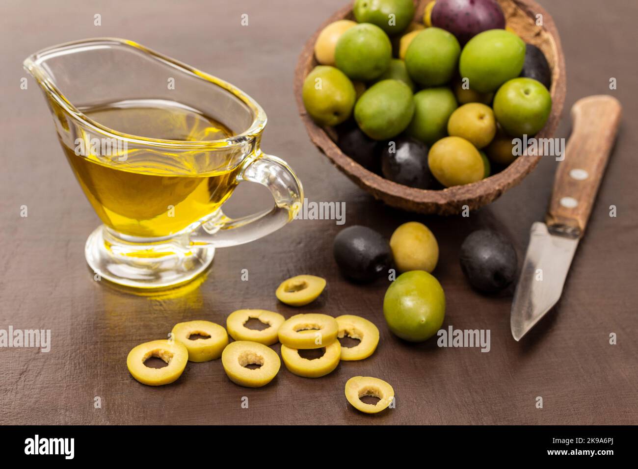 Olive verdi tagliate ad anelli e coltello sulla tavola. Olive di varie varietà in conchiglia di cocco. Olio d'oliva nel recipiente. Vista dall'alto. Sfondo marrone. Foto Stock