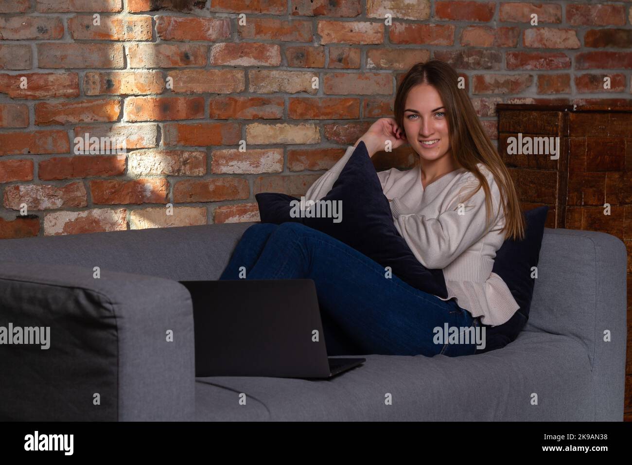 Donna sorridente in un maglione bianco seduto sul divano con un notebook davanti a lei Foto Stock