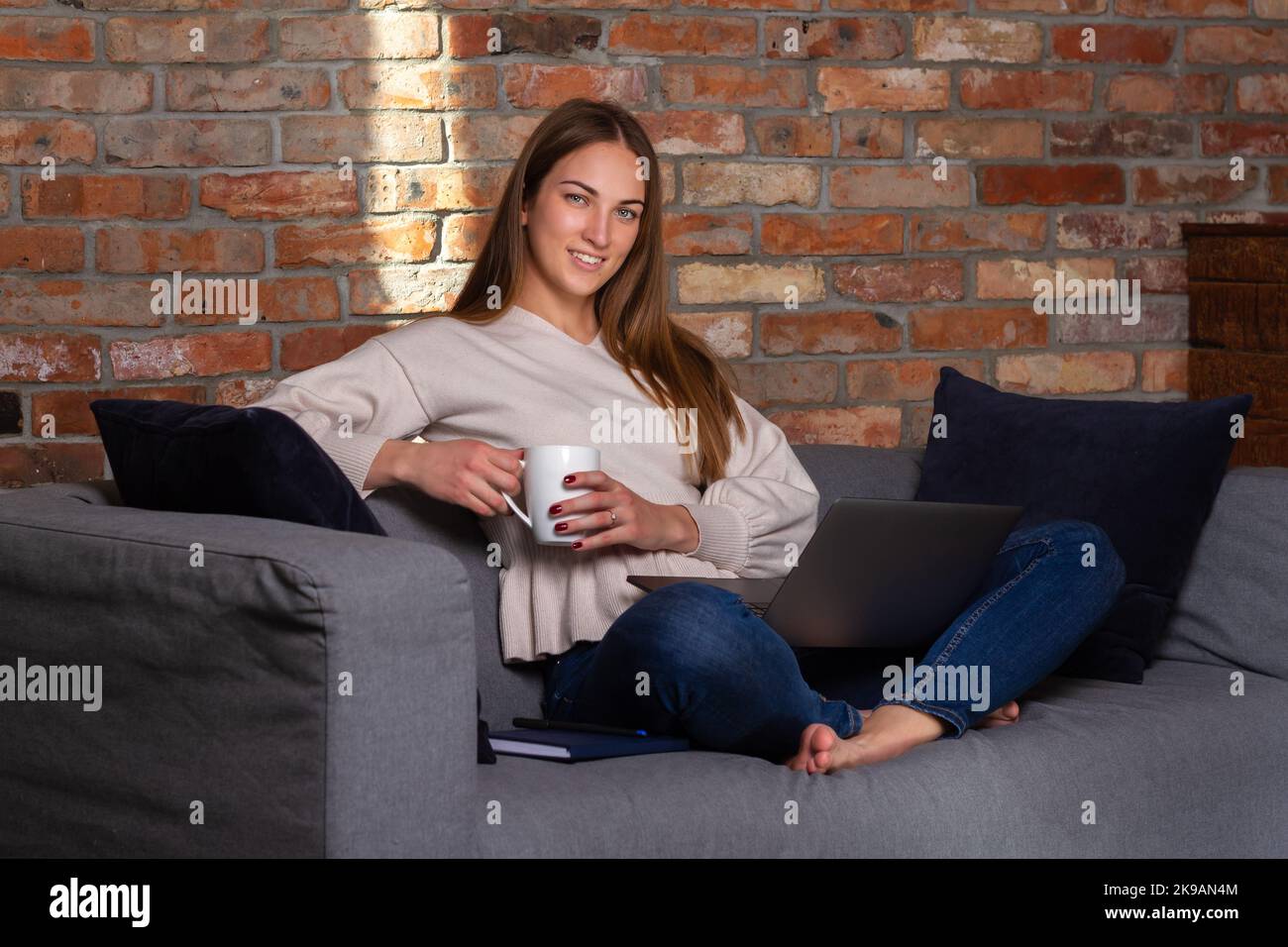 Donna sorridente in un maglione bianco che tiene una tazza bianca con un laptop in grembo e un notebook a lato Foto Stock