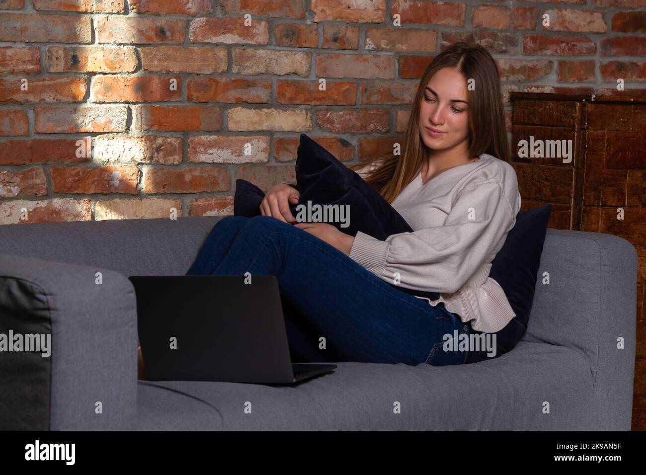 Donna sorridente in un maglione bianco seduto sul divano, tenendo un cuscino e guardando un notebook Foto Stock