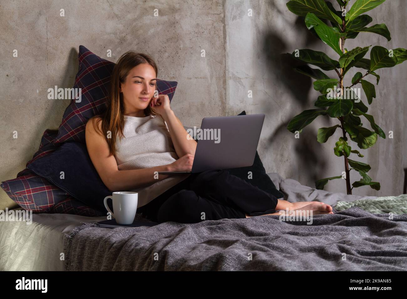 Donna in una parte superiore bianca seduta su un letto, guardando uno schermo di un notebook con una tazza bianca da parte Foto Stock