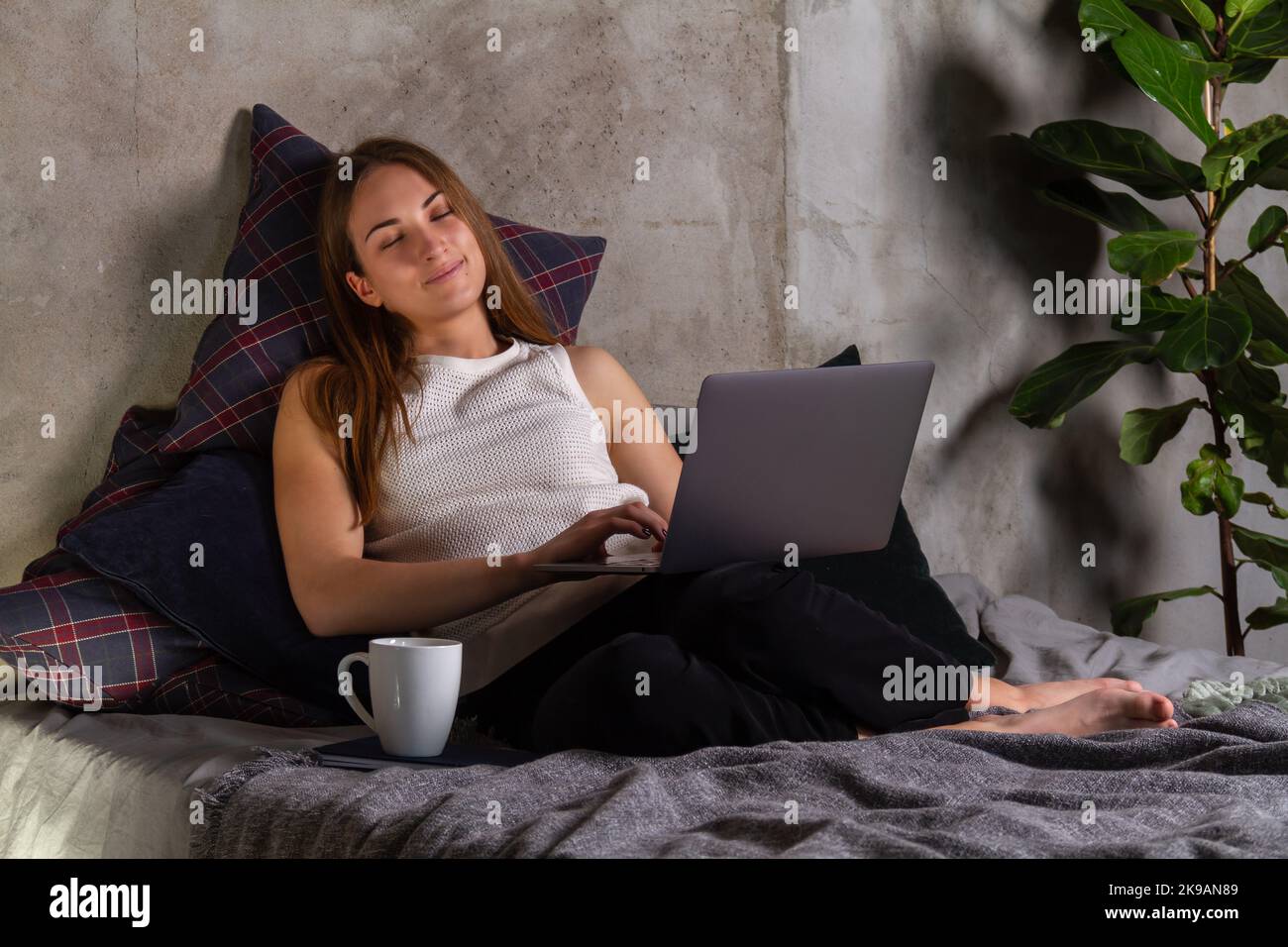 Donna addormentata in una parte superiore bianca che si rilassa sul letto con un laptop in grembo e una tazza bianca a parte Foto Stock