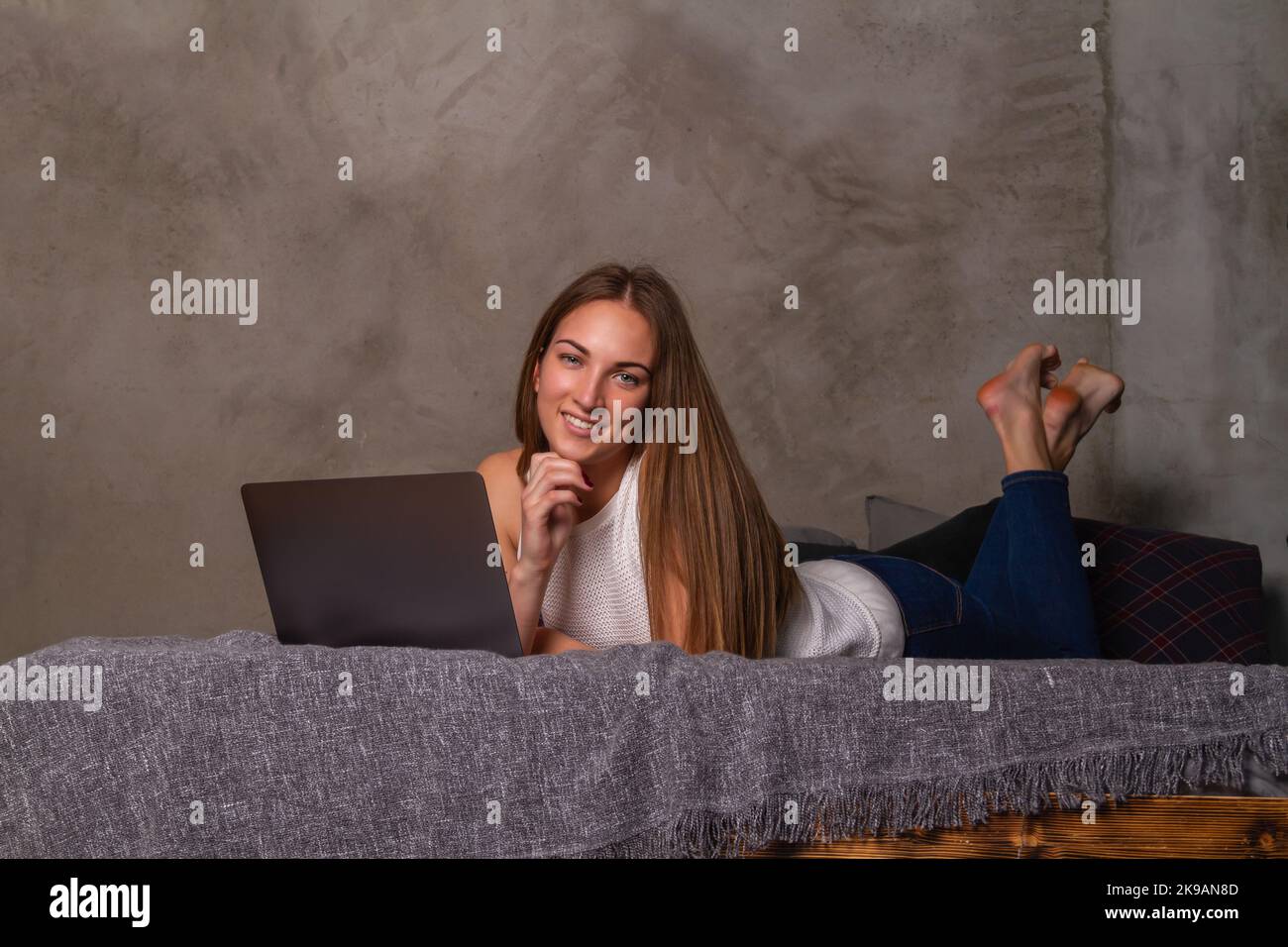 Donna sorridente in jeans e una parte superiore bianca sdraiata sullo stomaco a letto con un computer portatile davanti a lei e guardando nella macchina fotografica Foto Stock