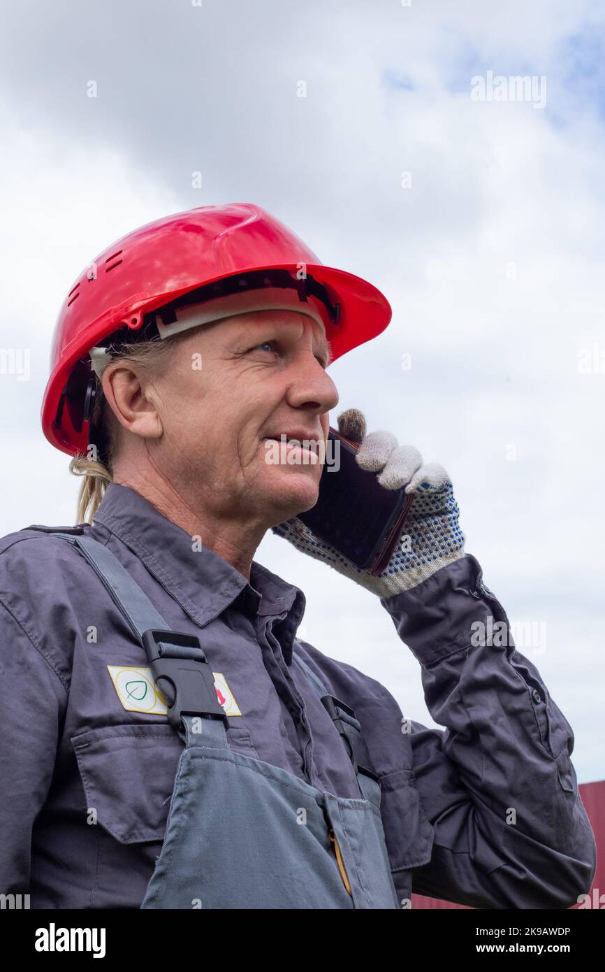 Un lavoratore adulto maschio in tuta e un elmetto sta parlando al telefono contro il cielo. Risoluzione dei problemi di lavoro. Foto Stock