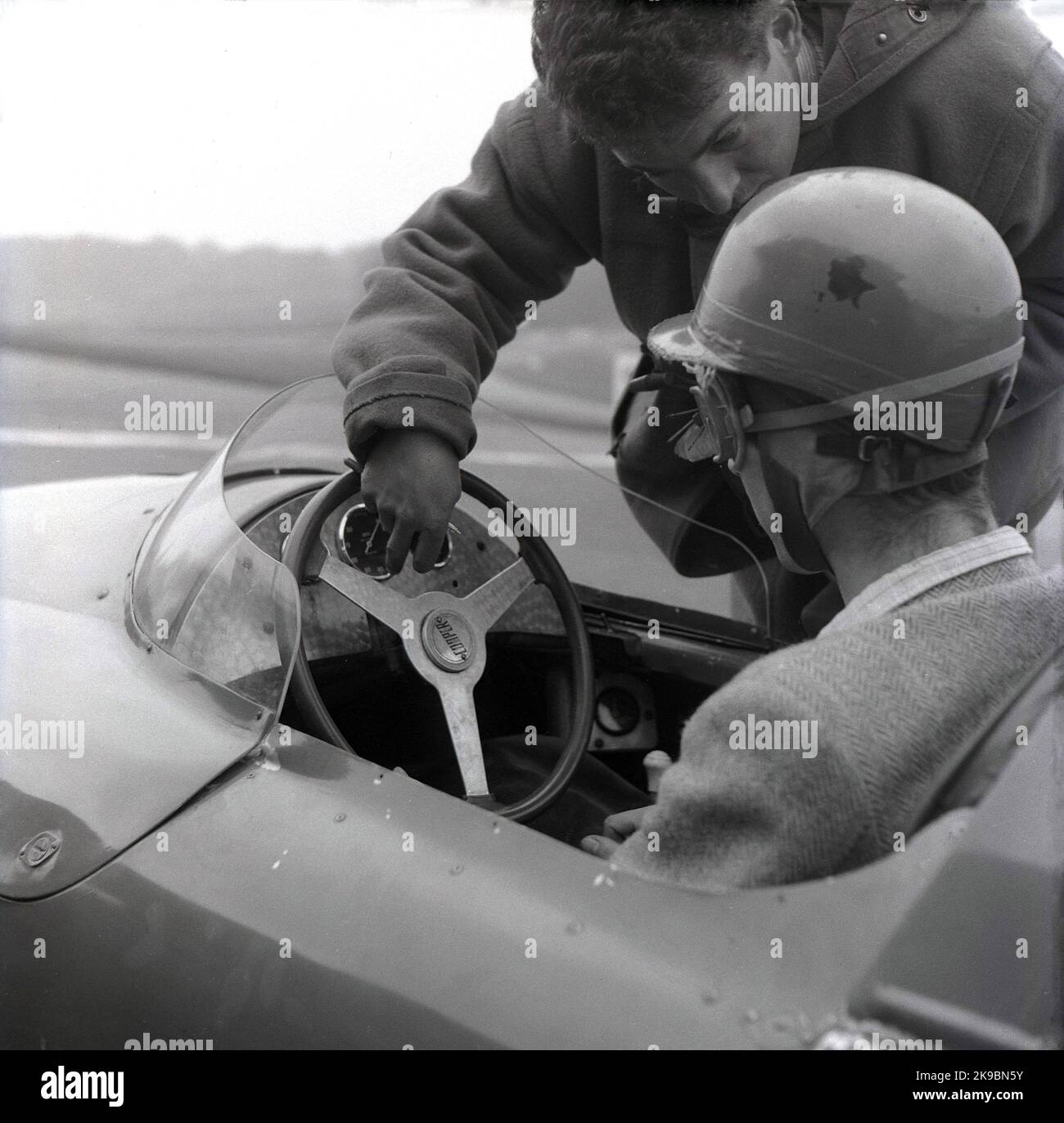 1959, storico, corse automobilistiche, scuola di guida. L'immagine mostra un istruttore maschio che parla con un pilota seduto in una vettura Cooper in pista al circuito automobilistico Brands Hatch, Kent, Inghilterra, Regno Unito. Il conducente indossa una giacca sportiva e un casco e googles dell'epoca. A questo punto, la Coopers Driving School si trovava in pista. Foto Stock