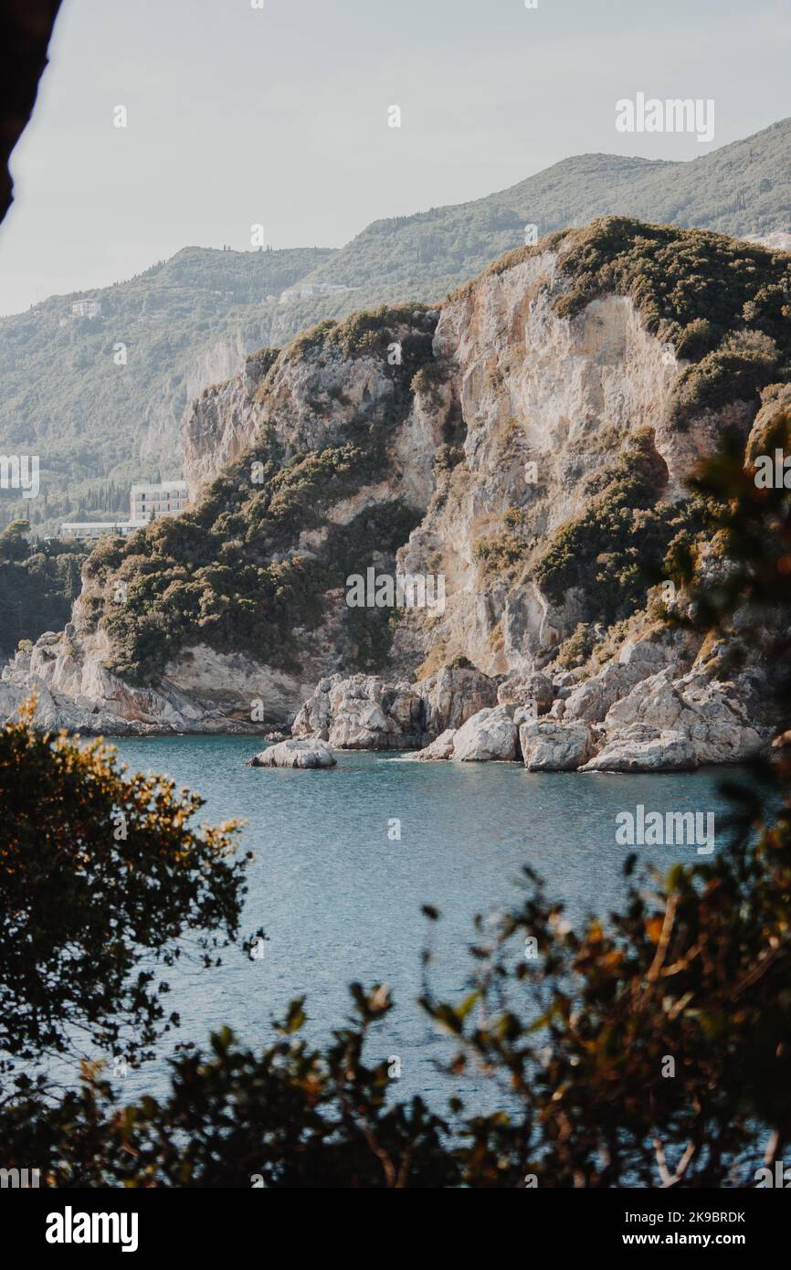Corfù, Grecia. Roccia grigia di montagna su cui crescono alberi verdi e piante dall'acqua blu. Vista attraverso l'albero nella foglia Foto Stock