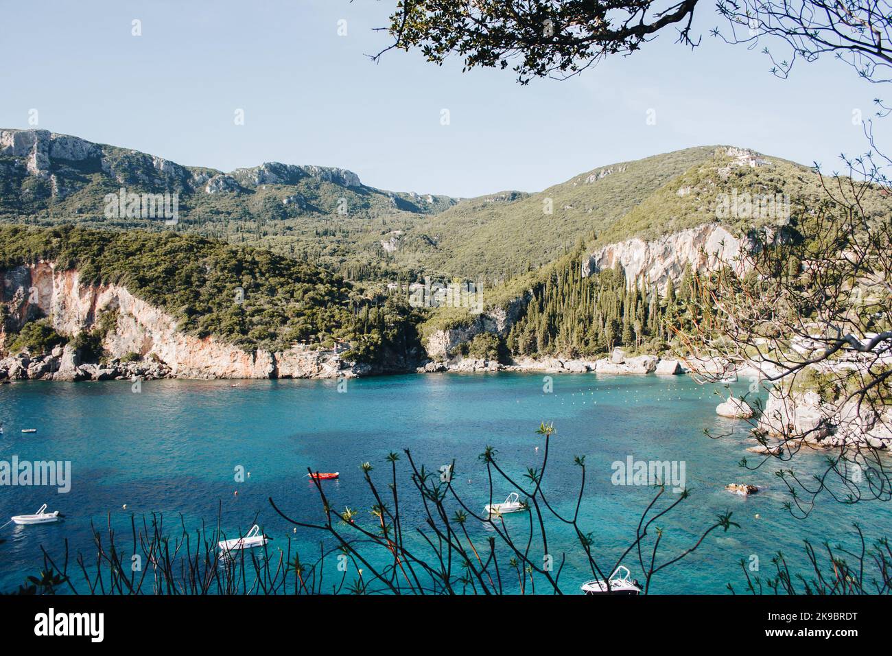 Corfù, Grecia. Una vista di un'enorme roccia di montagna con piante verdi e alberi che crescono su di essa accanto all'acqua blu dove si trovano le barche a motore. Foto Stock