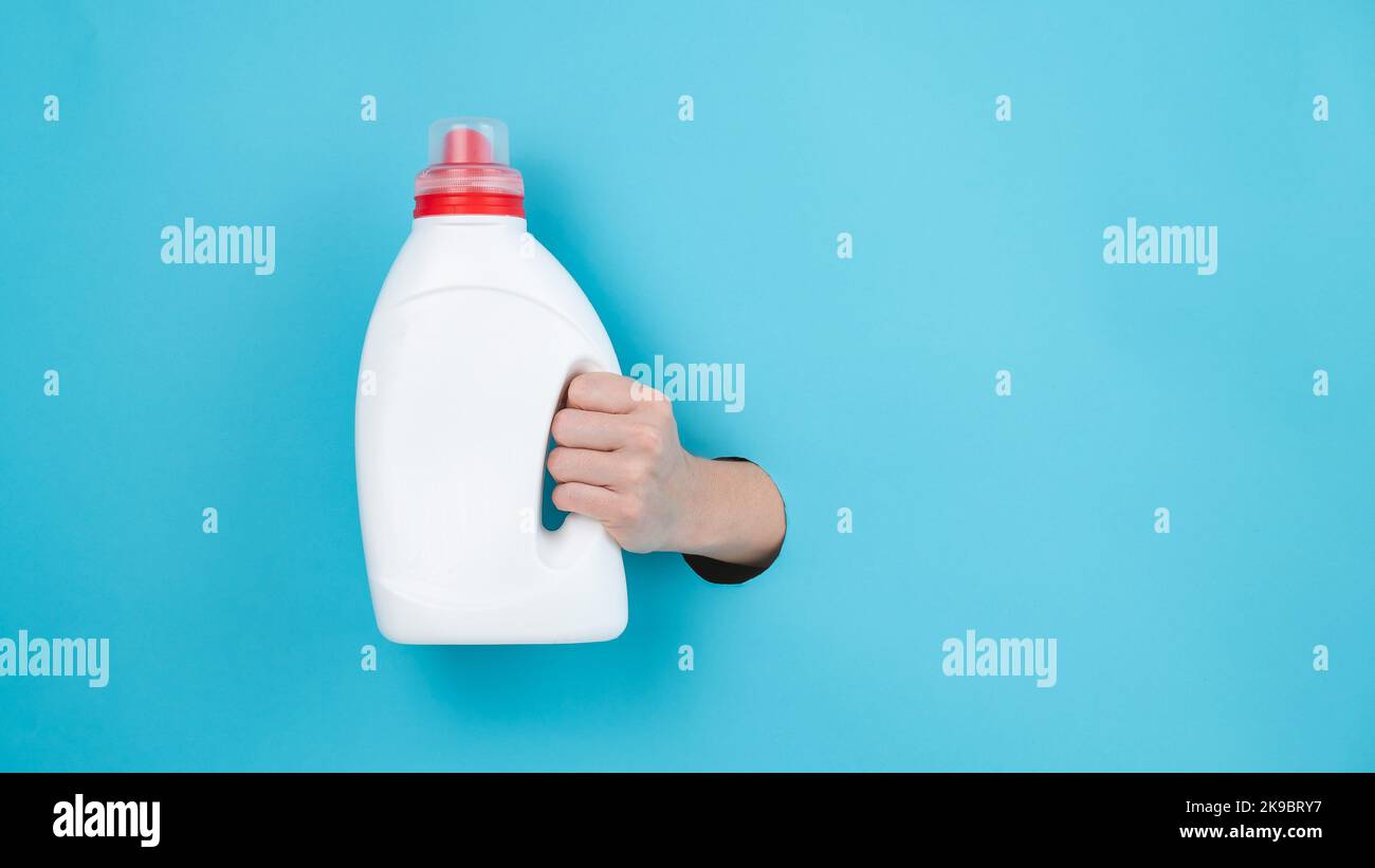 Una donna tiene una bottiglia bianca di detergente. Mano della donna che sporge da sfondo di carta blu. Foto Stock