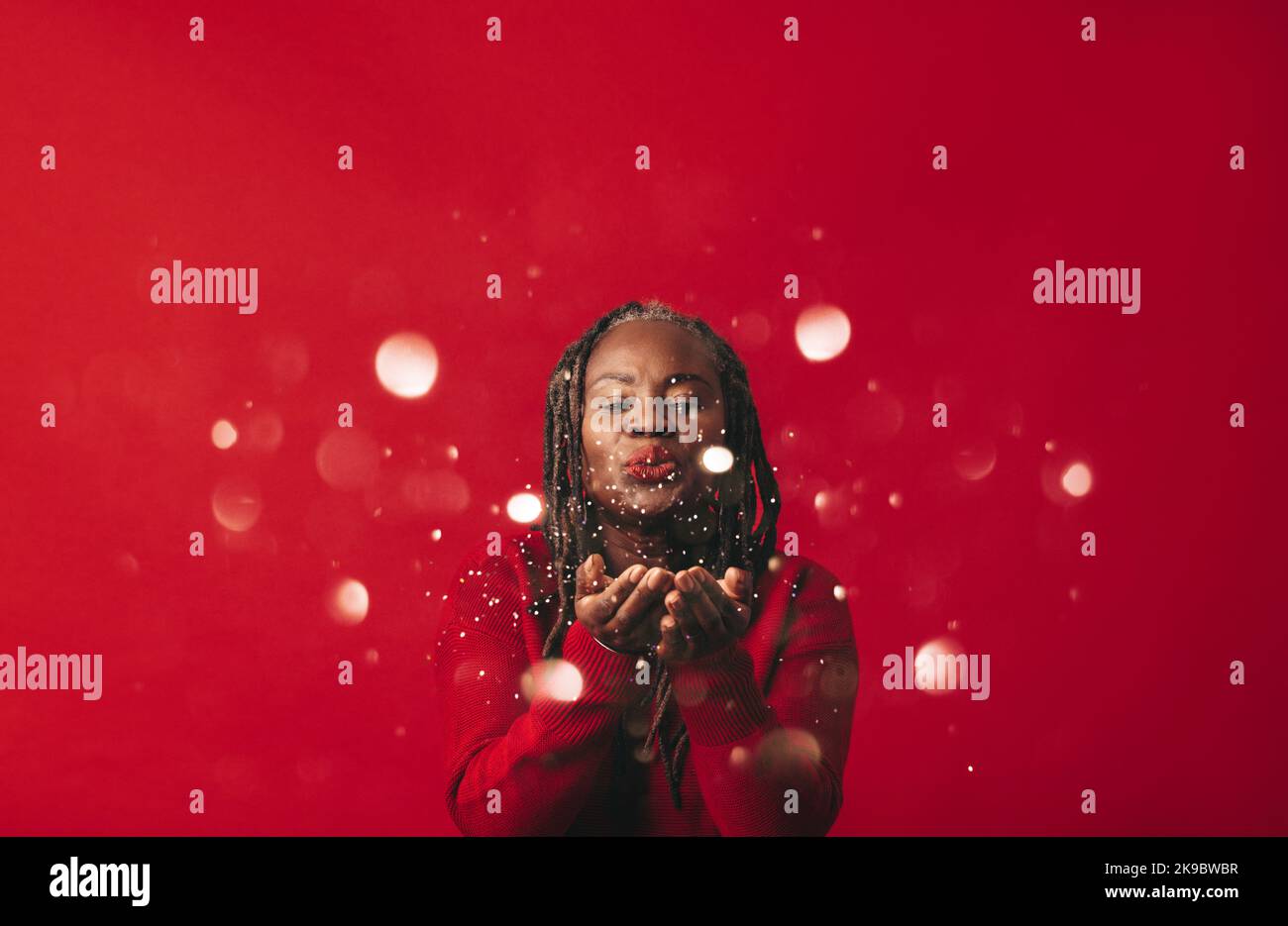 Felice donna nera che soffia i confetti nell'aria mentre si trova su uno sfondo rosso. Donna matura con Dreadlocks celebrare e divertirsi in un Foto Stock