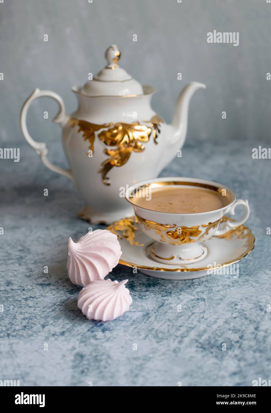 Raffinata tazza da caffè bianca e dorata su sfondo blu astratto. Caffè con un'appetitosa schiuma dorata. Due meringhe ricce rosa accanto alla tazza. B Foto Stock