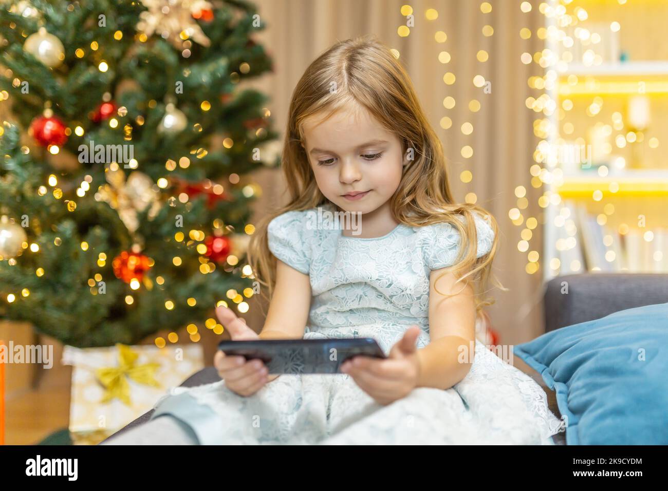 Una bambina sta guardando un video su uno smartphone per Natale, sua figlia sta celebrando il nuovo anno online vicino all'albero di Natale Foto Stock