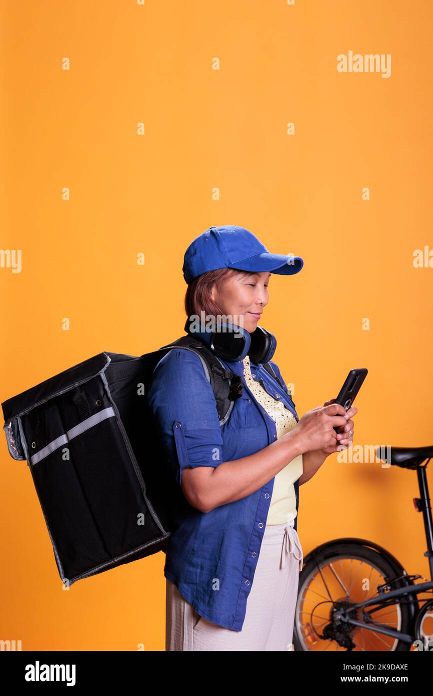 Donna anziana consegna uno zaino di cibo da portare via controllando le istruzioni e i dettagli dell'ordine sullo smartphone. Donna con consegna uniforme utilizzando la bicicletta come trasporto. Servizio di ristorazione a casa Foto Stock