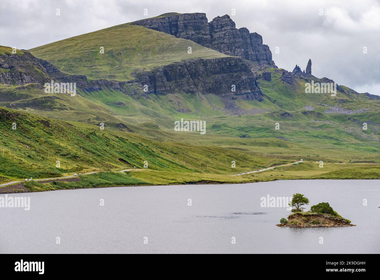 Il Storr, oltre il lago, in lontananza, un prominente punto di riferimento Skye, scogliera frastagliata pareti scogliera, piccola isola di roccia che sporge dall'acqua, strada di montagna ne Foto Stock