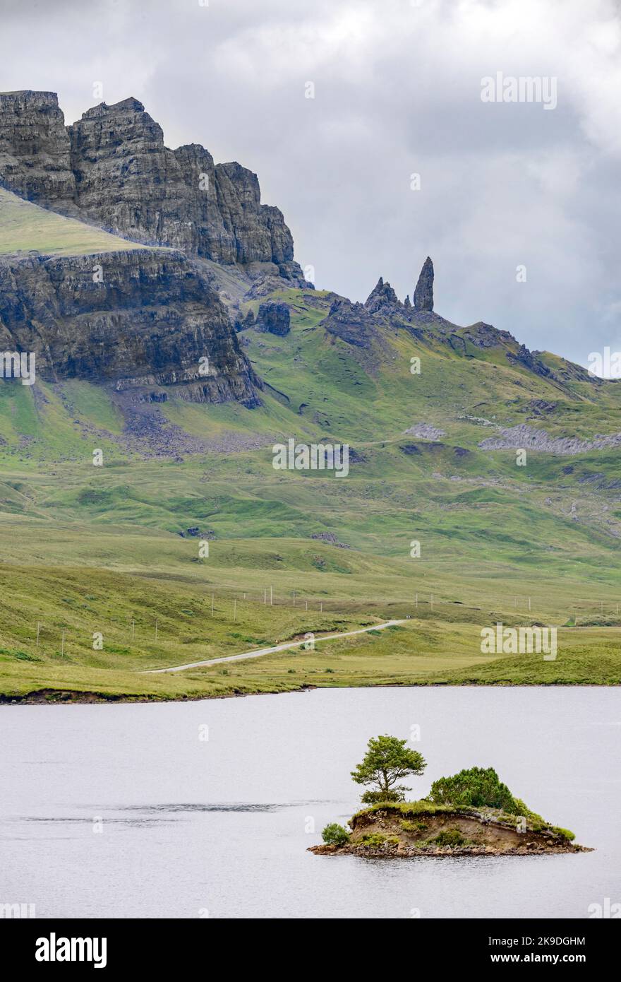 Il Storr, oltre il lago, in lontananza, un prominente punto di riferimento Skye, scogliera frastagliata pareti scogliera, piccola isola di roccia che sporge dall'acqua, strada di montagna ne Foto Stock