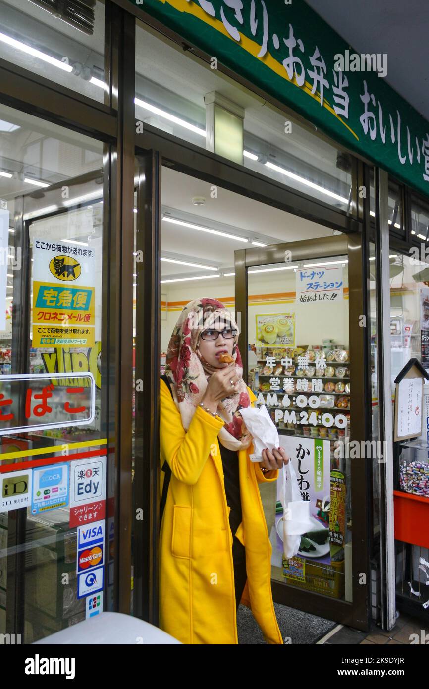 Ritratto di giovane donna musulmana che indossa hijab e occhiali in piedi e mangiare spuntini alla porta d'ingresso del minimarket. Foto Stock