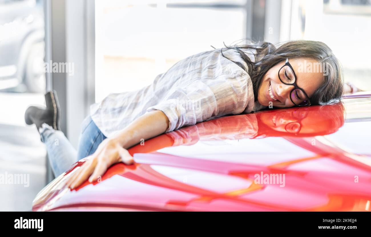 Donna eccitata che abbraccia la sua nuova auto rossa da sogno in una concessionaria di auto. Foto Stock
