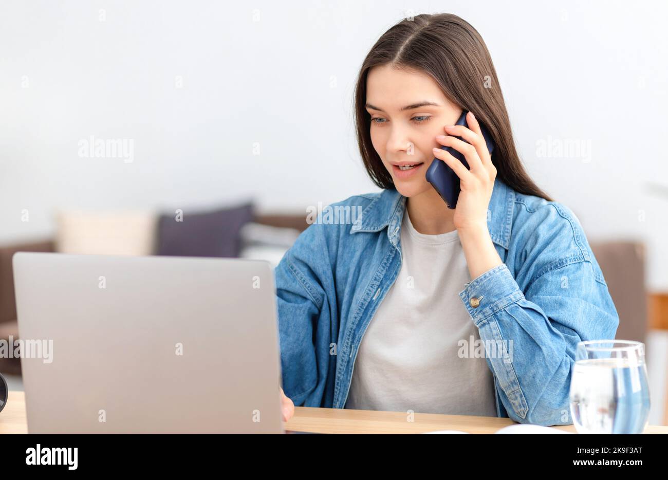 Giovane donna caucasica freelance o dipendente in ufficio che lavora in remoto utilizzando un computer portatile e parlando con un telefono cellulare Foto Stock