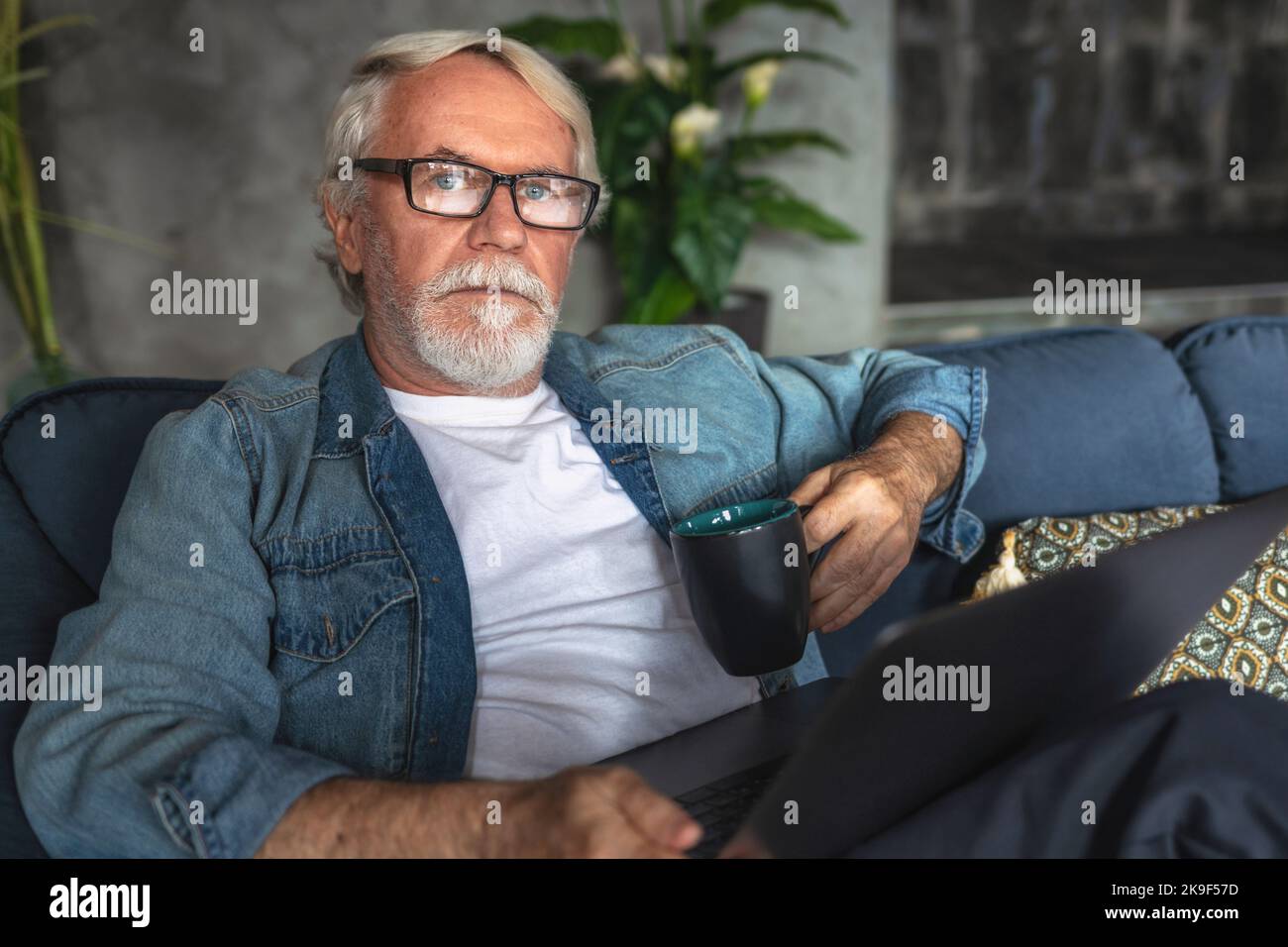 Ritratto di un uomo d'affari anziano con una tazza di caffè e un computer portatile seduti sul divano a casa o in un ufficio moderno Foto Stock