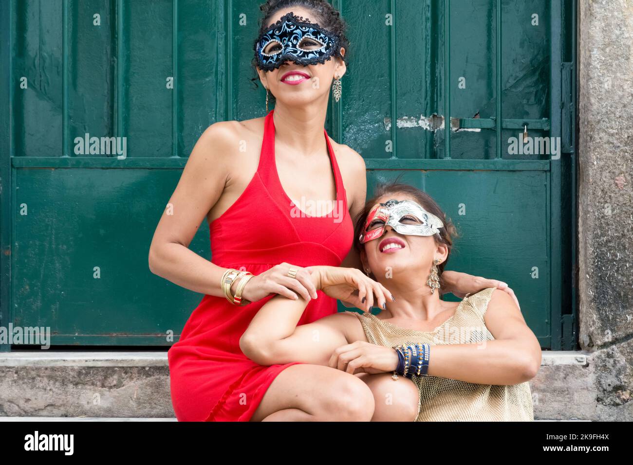 Due ragazze che indossano la maschera di carnevale nei vestiti estivi chiari stanno sedendosi contro una porta di colore verde. Ragazze di città lontano dalla capitale. Foto Stock