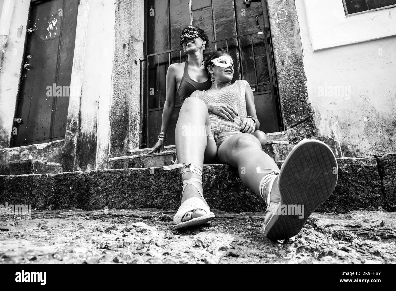 Vista bassa in bianco e nero di due ragazze che indossano una maschera di carnevale seduta contro una porta di colore verde. Ragazze di città lontano dalla capitale. Foto Stock