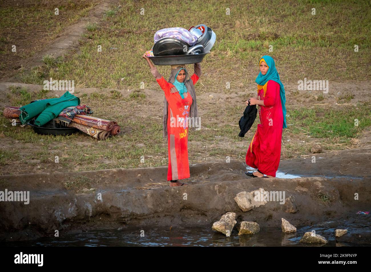 Due donne egiziane, una che porta le pentole di metallo sulla testa, vestite di rosso brillante, pentole di lavaggio, padelle e tappeti sulle rive del fiume Nilo, Egitto Foto Stock