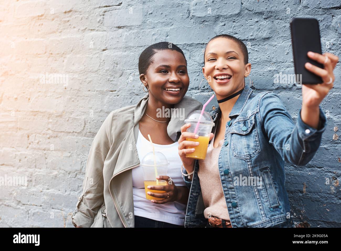 Chiacchiera dal vivo ovunque e in qualsiasi momento. Due giovani donne in piedi accanto a un edificio sorridendo e scattando selfie mentre sorreggono le loro bevande fresche. Foto Stock