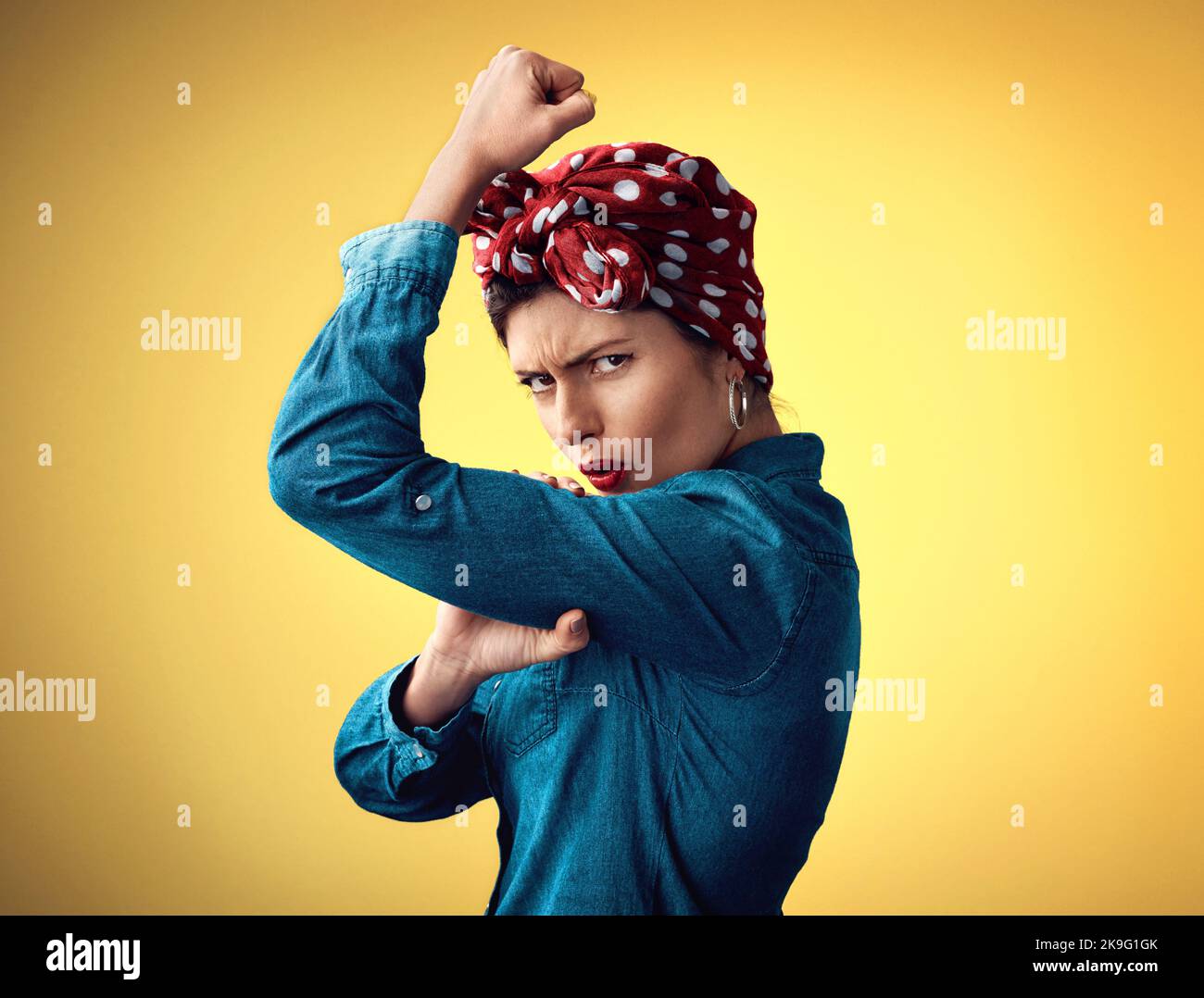 Forte, all'interno e all'esterno. Studio ritratto di una giovane donna attraente flexing il suo bicep mentre si trova su uno sfondo giallo. Foto Stock