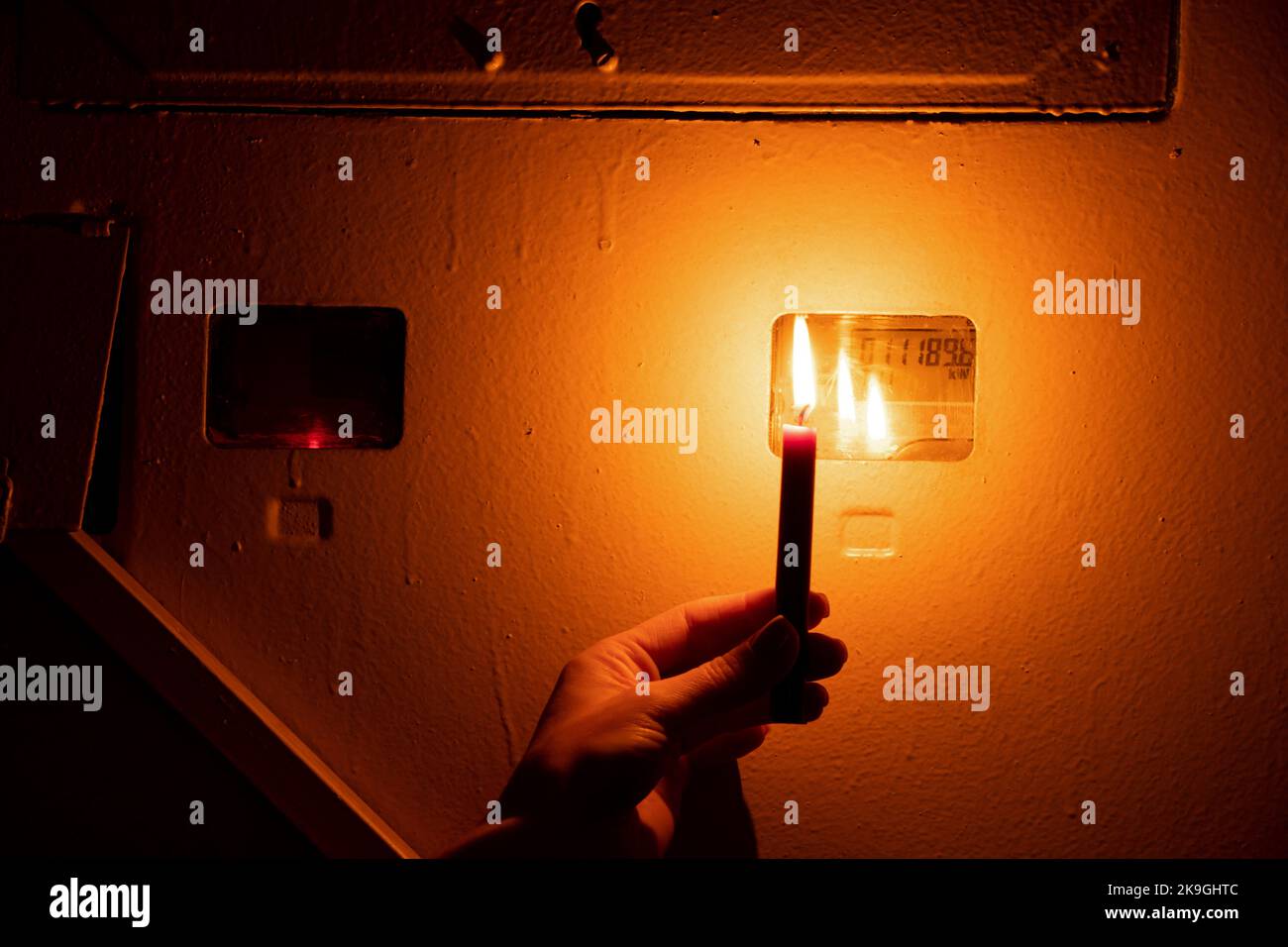 La mano di una donna tiene una candela accanto a uno scudo con un metro in un appartamento, Ucraina senza elettricità a causa di guerra, interruzioni di corrente Foto Stock